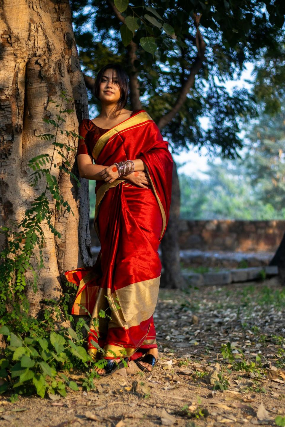 A woman in a red and gold sari standing next to a tree